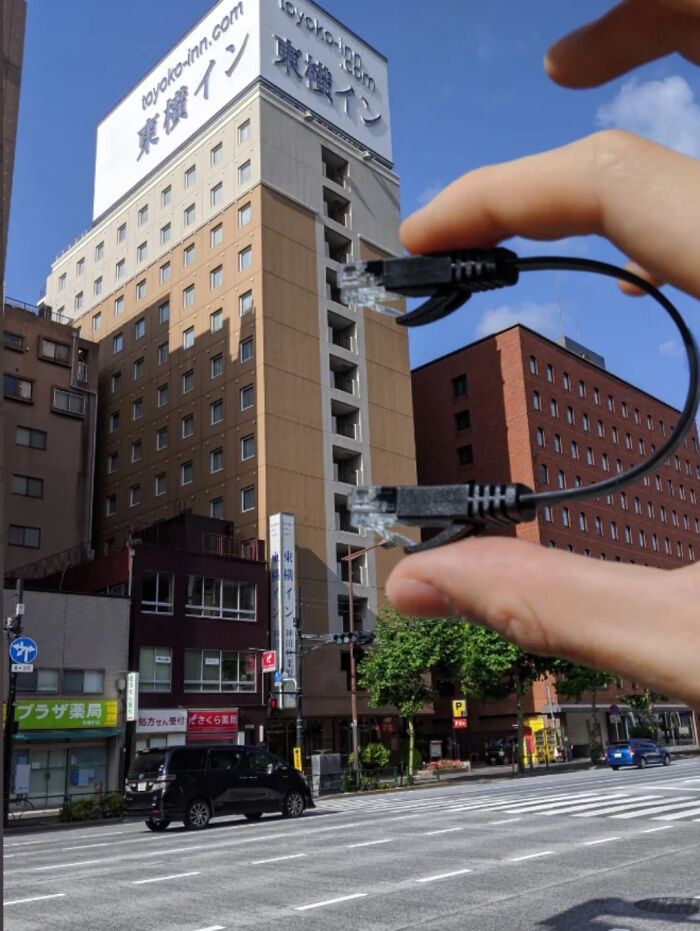 Fingers holding cable above a city street, resembling a giant plug for the building, creating a quirky optical illusion. Fingers holding cable above a city street, resembling a giant plug for the building, creating a quirky optical illusion.